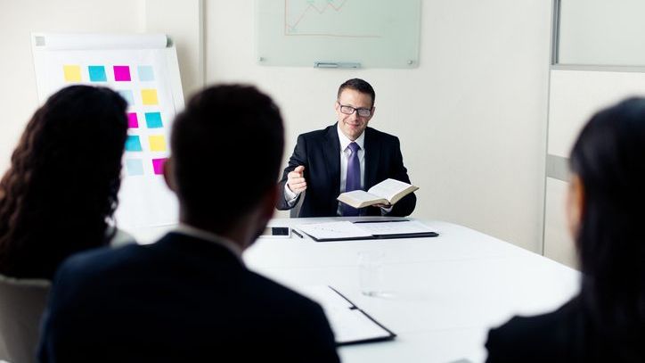 A man in a suit holding a Bible and speaking with others in a conference room.