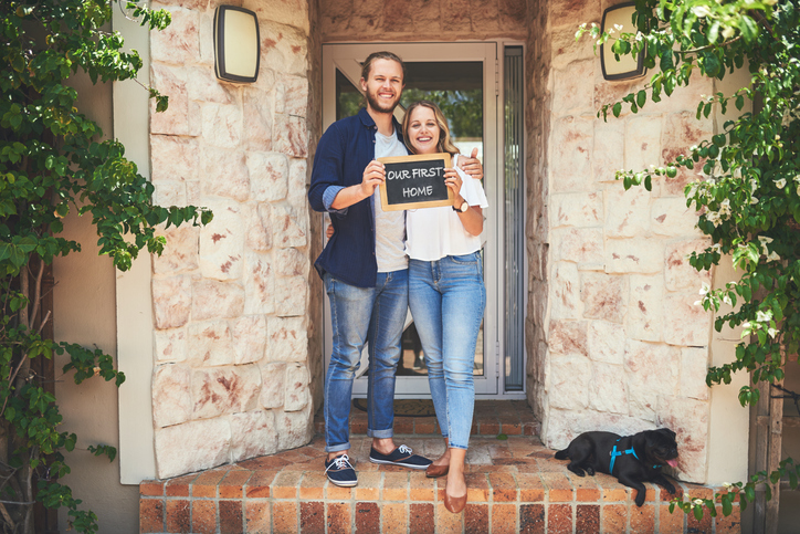 A couple pose in front of their new home, their first real estate purchase.