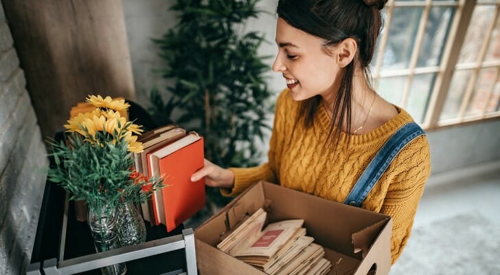 Image shows a young adult standing in their new apartment, arranging some books on a bookshelf in their apartment. SmartAsset analyzed data on rent, housing units and earnings to conduct this year's study on the cities where renters can afford to live alone.