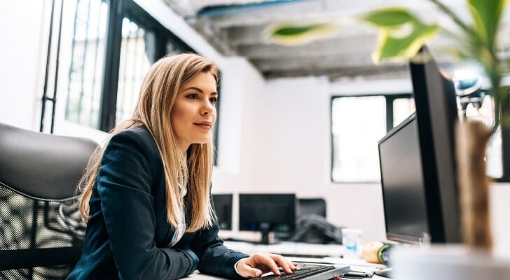 Image shows a young woman financial advisor wearing a black suit sitting at a desk and working at a computer station. In this study, SmartAsset identified the best cities for young financial advisors to start their careers.