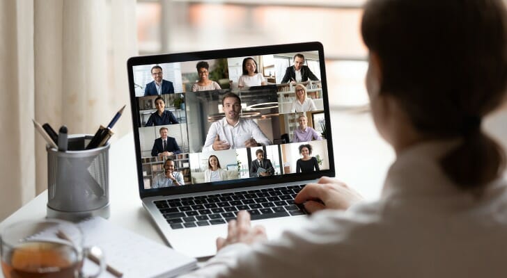 A young worker wearing a beige shirt and ponytail sits at a desk with their back facing the viewer; we see the view over their shoulder of their desk and laptop screen filled with faces of colleagues on a video call. SmartAsset analyzed various data sources to find the fastest-growing job for young professionals.