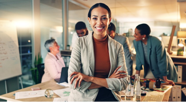 A businesswoman poses while colleagues work around a conference table in the background.