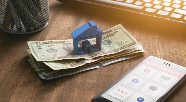 Image shows a blue house figurine on top of a stack of a few bills; these sit on a desk, next to a smartphone that shows the interface of a budgeting app. SmartAsset analyzed Census data to conduct this study on the hours of work needed to pay rent in the 25 largest U.S. cities.