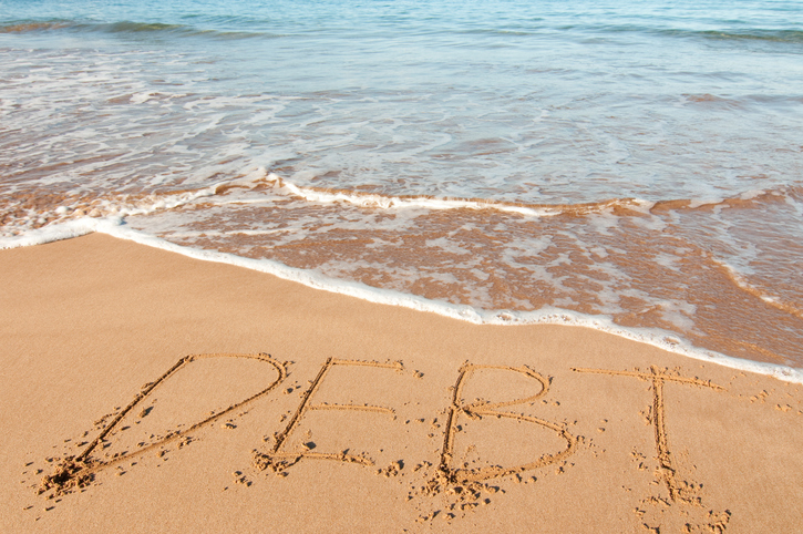 "Debt" written in the sand. When a deceased spouse leaves debt behind, it’s possible that debt collectors could attempt to sue you to recover what’s owed.