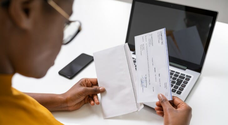 Image shows a woman who is wearing glasses sitting at a desk in front of a laptop and removing a paycheck from an envelope in her hands. SmartAsset analyzed data to find the best cities for women's pay.