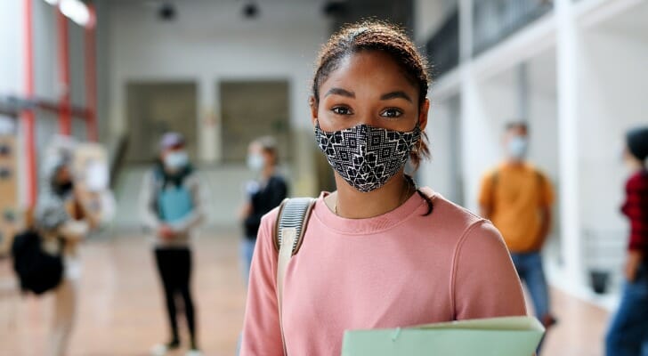Image shows a student standing in a school hallway, holding notebooks and wearing a face mask to protect from COVID-19 and other viruses. SmartAsset analyzed various data sources to conduct its latest study on the top U.S. states for higher education.