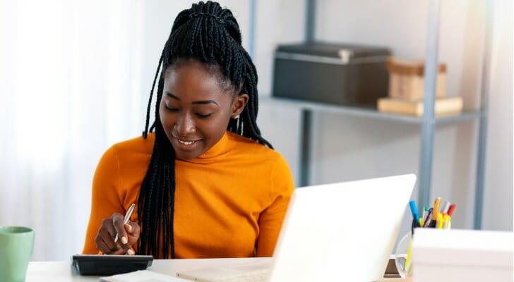 Image shows a person sitting at a table using a laptop and calculator to understand what investing-related fees they might incur.