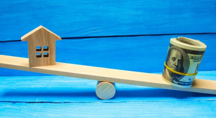 Tiny seesaw with model house on one end and a wad of bills on the other