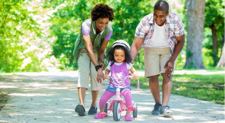 A mother and father teacher their daughter how to ride a bike in a park.