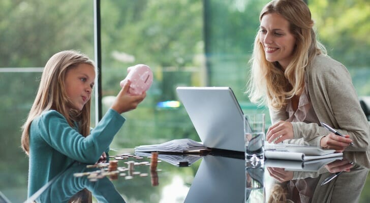 Image shows a parent and a child sitting at a table and talking about how much money is in the child's piggy bank.