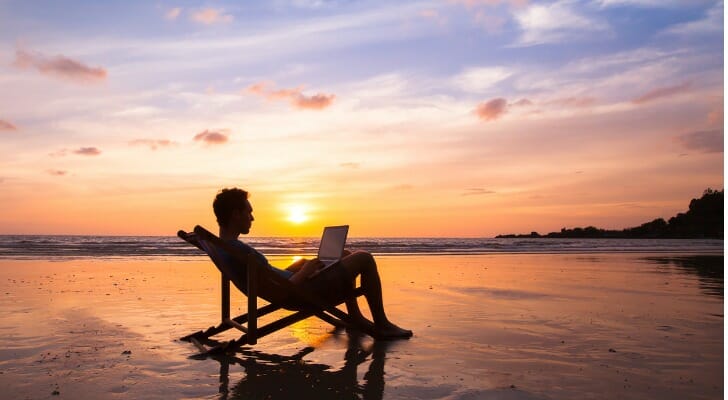 A man relaxes on the beach, reviewing offshore bank accounts on his laptop.