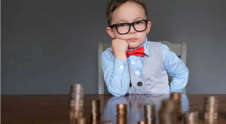 A boy with glasses and a red bowtie sitting behind stacks of coins on a table.