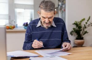 Senior man sits at table looking at papers with a pen in hand.