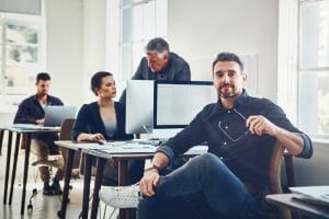 A man looks at the camera while sitting at his desk with computer. Other people work in the room behind him.