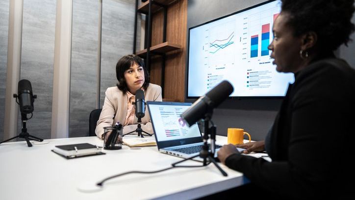 Two woman record a podcast in front of a large screen showing charts and data.