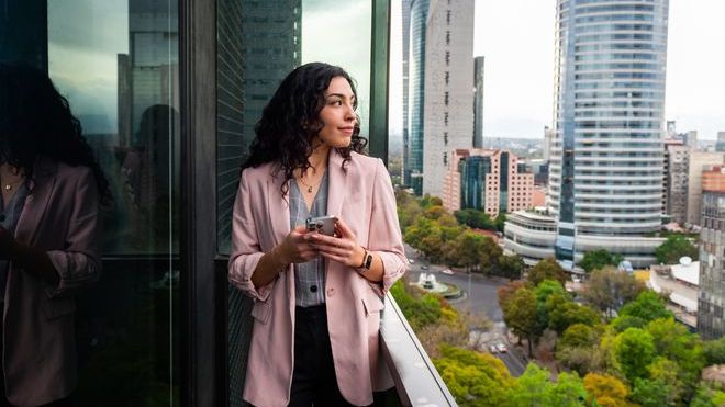 A wealth management looks out onto the city while checking her email on her phone.