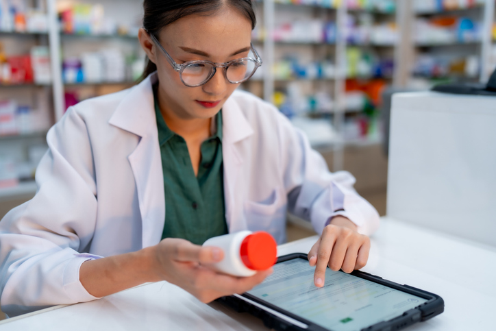 A pharmacist cross-checks medication details on a digital tablet while holding a prescription bottle.