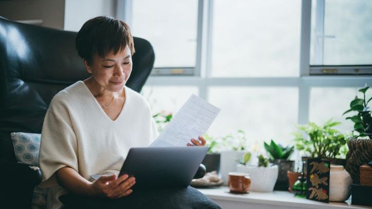 A woman looks over some paperwork while resting a laptop in her lap.