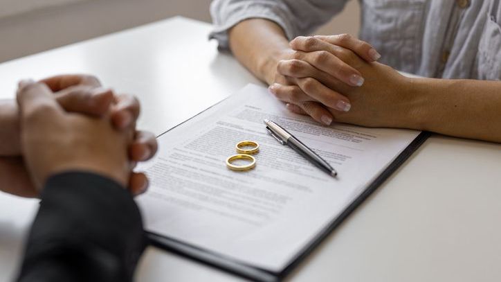 Two people sit across from each other with divorce paperwork and wedding rings between them.