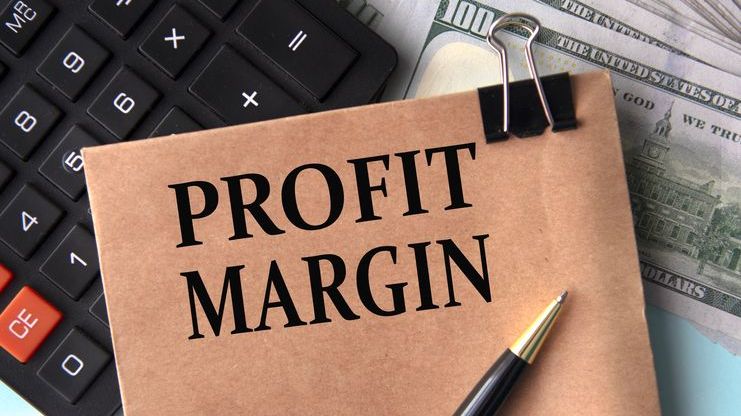 A brown envelope with the words "profit margin" printed on it rests on a calculator and banknotes.