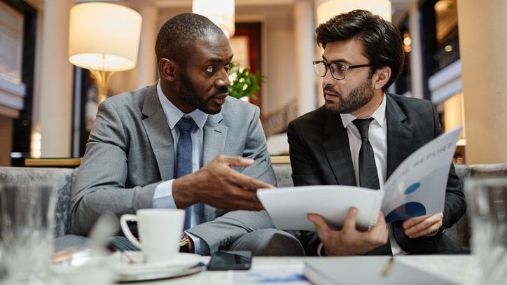 Two businessmen discuss a deal while sitting at table in hotel lobby.