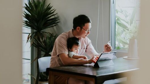 A young girl sits on her dad's lap while he works on his tablet.