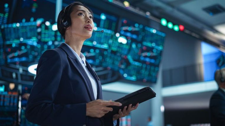 A female broker wears a headset and holds a tablet on the floor of a stock exchange.