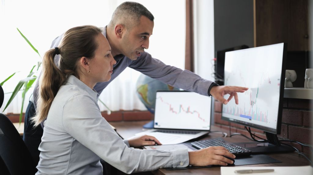 Colleagues review stock price charts on a pair of monitors in an office setting.