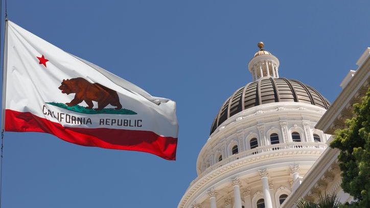 The California state flag waves in the wind in front of the state capitol.