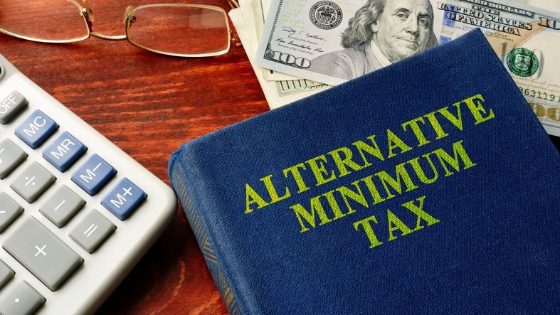 A book with the title "Alternative Minimum Tax (AMT)" rests on a desk next to a keyboard, glasses and cash.