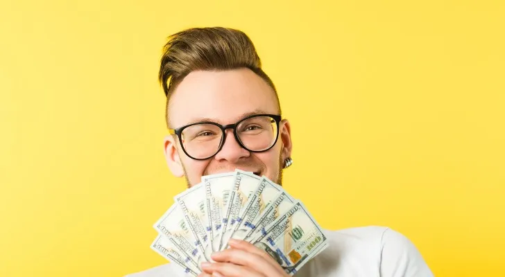 A man in his 20s or 30s smiles while holding $100 bills in front of his face.