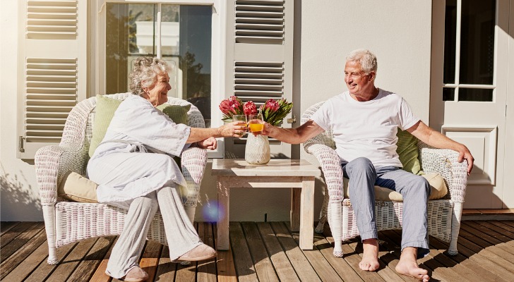 A man and woman who appear to be in their 70s toast while enjoying beverages together on their deck.