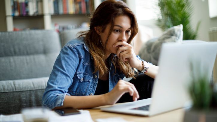 A woman has an anxious look on her face as she browses on her laptop while sitting at her coffee table.