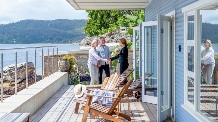 A couple shakes hands with a real estate agent on the deck of a waterfront home.