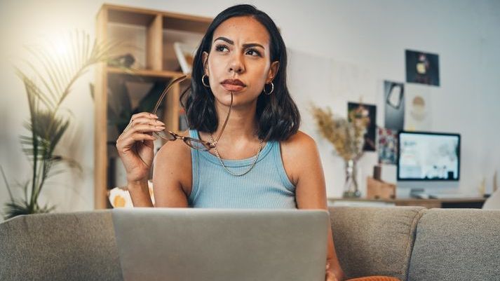 A woman contemplates while sitting on a couch with a laptop computer.