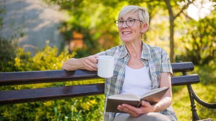 A senior woman enjoys reading a book and drinking coffee on a bench in her garden.