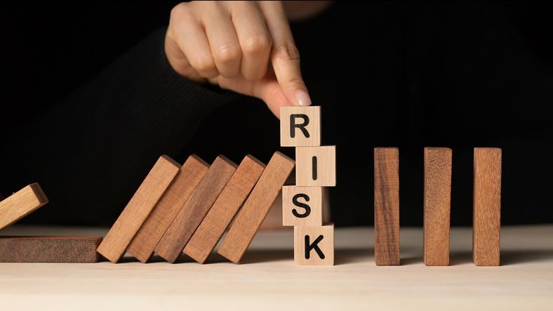 A close-up of a woman's hand balancing wooden blocks that spell "RISK," while dominos fall in the background.