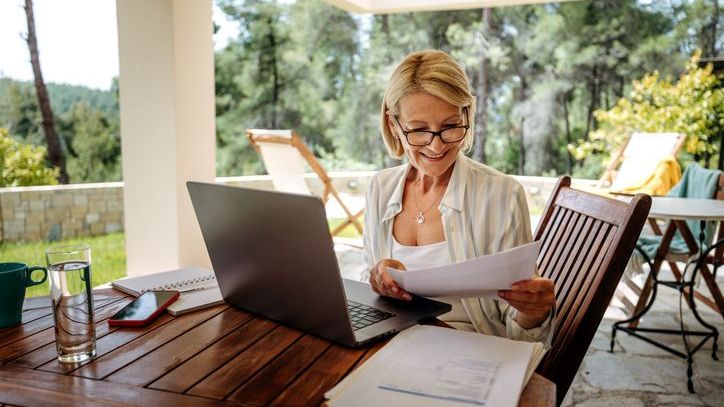 A smiling mature woman working on a laptop on the back patio of her home.