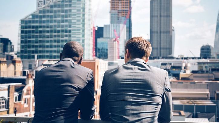 A rear-facing view of two businessmen standing on a rooftop looking out over a cityscape.