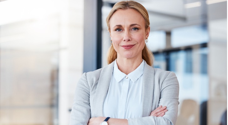 A woman wearing business attire stands in an office setting with her arms crossed.