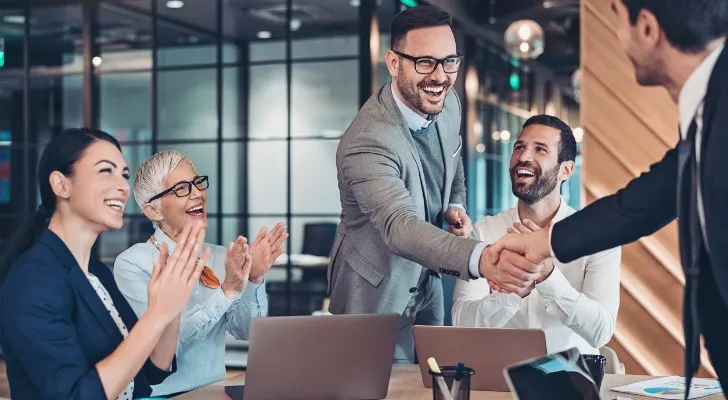 Two men in suits shake hands at a conference table while colleagues applaud.