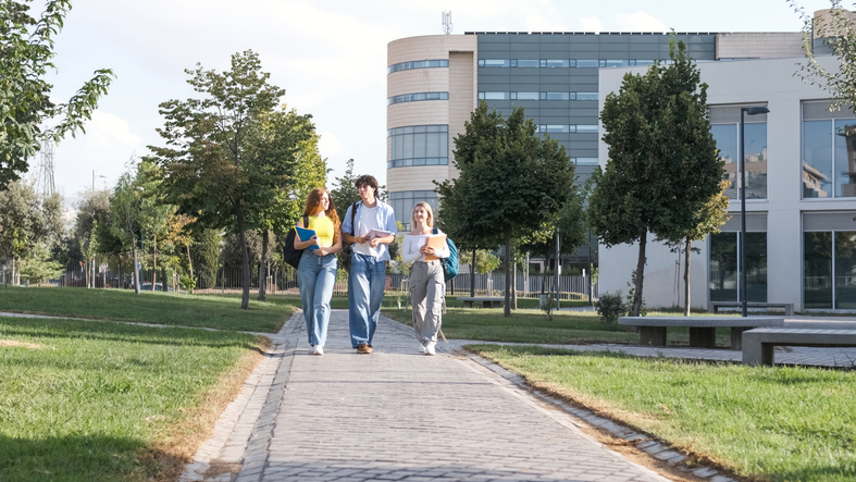 Full body of young man with female friends in casual clothes chatting while walking on university campus footpath in city on sunny day