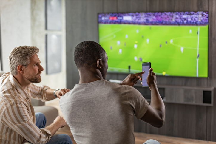 Two friends holding smartphone with sports bets app while watching football match at home in living room.